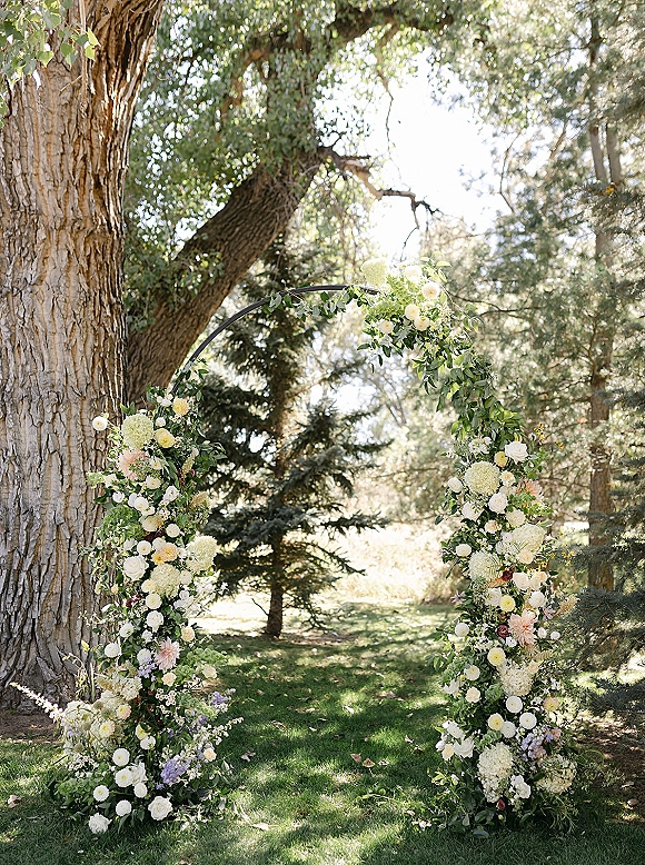 Wedding ceremony arch with asymmetrical white, blush, and lavender flowers on a metal frame, set on a sunny garden lawn under evergreens