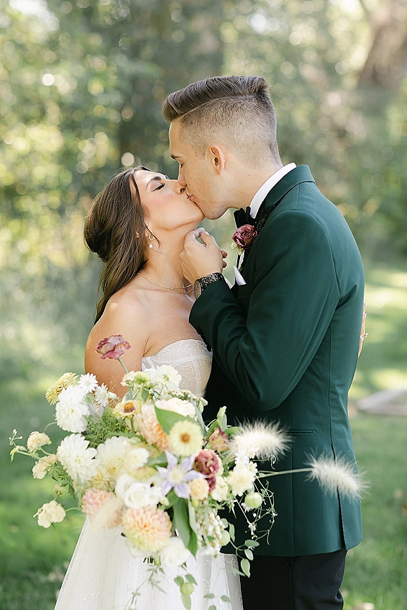 Wedding kiss portrait of bride and groom kiss, her strapless lace dress and pastel bouquet against soft greenery and trees in natural light