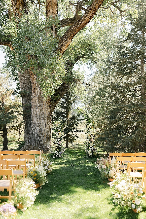 Outdoor ceremony setup with wood folding chairs lining a flower-marked aisle, floral pillars and greenery on a sunlit lawn under a large tree