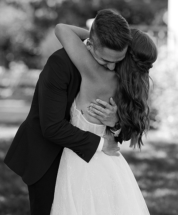 Wedding couple portrait of groom in black suit hugging bride in a lace open-back gown on a garden path with blurred greenery