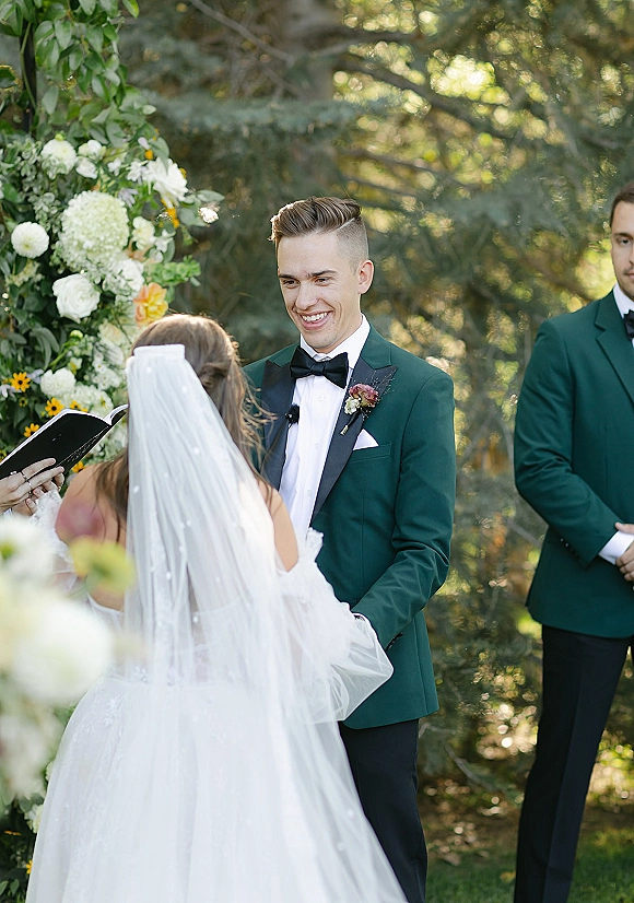 Wedding vows as bride and groom hold hands under a floral ceremony arch, bride’s veil trailing and officiant reading in a garden setting