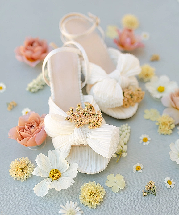 Bridal shoes in a wedding shoe flat lay with white bow heels, ankle straps, pleated fabric and floral appliques on light blue surface