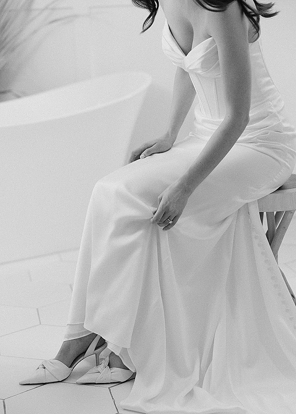 Bridal dress in strapless satin wedding dress with corset bodice, wedding ring and pointed toe heels beside a wooden stool on tile floor