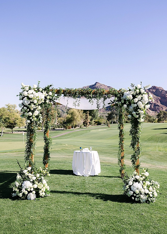 Wedding ceremony arch with white roses and greenery garland on a wooden arbor, sheer drape and table set on a mountain-view lawn