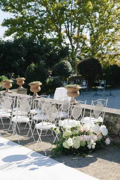 Ceremony seating with outdoor wedding ceremony chairs lining a white aisle runner on gravel, with hydrangea and rose florals at the entrance on a stone terrace