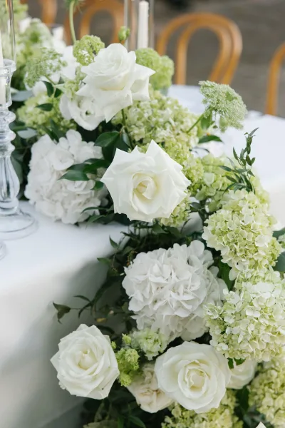 Wedding table flowers with a white rose centerpiece and hydrangeas, lush greenery runner, and clear glass candlesticks on a white tablecloth in a reception space