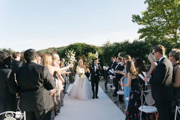 Ceremony recessional as bride and groom walking aisle past white chairs, guests tossing confetti petals beneath a floral arch in sunny garden