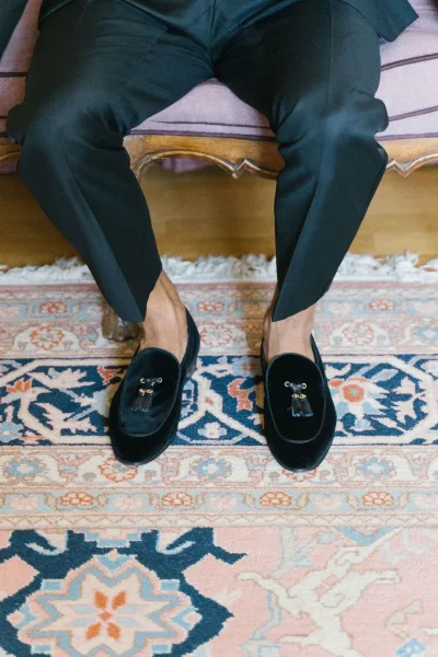 Groom shoes in black velvet loafers with tassels beside suit trousers, resting on an upholstered bench over a vintage rug