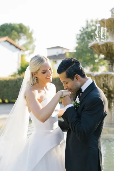 Wedding couple portrait of groom kissing bride’s hand as they hold hands, her veil flowing beside a fountain with trees and building behind