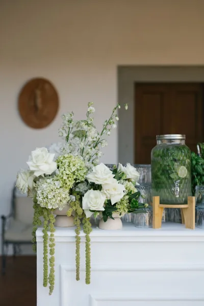 Wedding cocktail station with white floral arrangements, ceramic vases, and a glass dispenser of lemon mint water on a white bar counter indoors