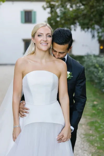 Couple portrait of bride and groom embrace as he hugs her from behind and kisses her forehead outside a white estate building