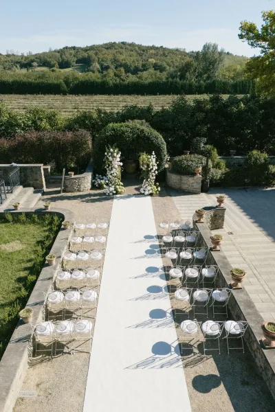 Outdoor ceremony setup with a white aisle runner leading to a floral arch, white chairs on a stone terrace with hills beyond