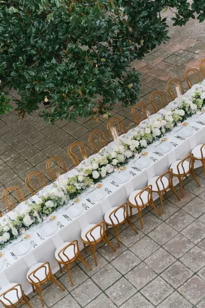 Reception tablescape with a long banquet table wedding setup, white floral and greenery runner, taper candles, and place settings on a stone patio under a leafy tree canopy