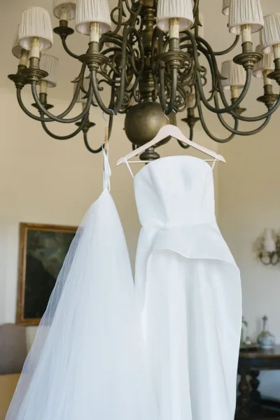 Wedding dress, a strapless bridal gown with tulle skirt and veil on wooden hangers beneath a chandelier in an indoor room