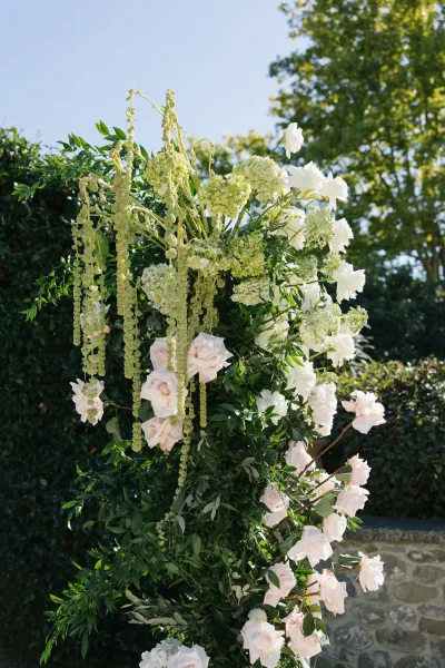 Wedding floral arrangement on a ceremony floral pillar with white and blush roses and hydrangea against garden trees and a stone wall