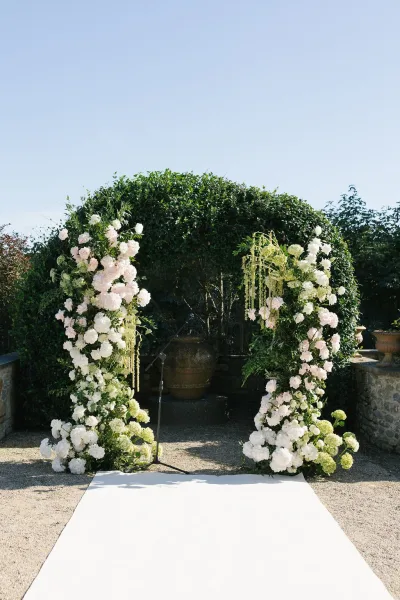 Wedding ceremony backdrop with a greenery wedding arch of white flowers and blush roses, hanging florals, and aisle runner in a garden courtyard