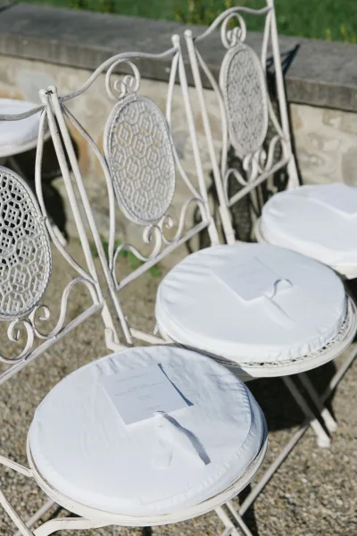 Ceremony seating with wedding ceremony chairs, white metal cushions and ribbon-tied programs on gravel in a stone-walled courtyard with greenery