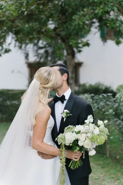 Wedding kiss portrait of bride and groom kissing, bride holding a white and green bouquet with veil flowing in a garden by a white building