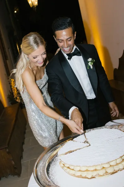Wedding cake cutting as bride in a sequined dress and groom in tuxedo slice a powdered sugar cake by lantern light outdoors at night