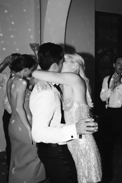 Wedding dance kiss as the bride in a strapless beaded dress and groom in a bow tie embrace under dance floor lighting with guests behind