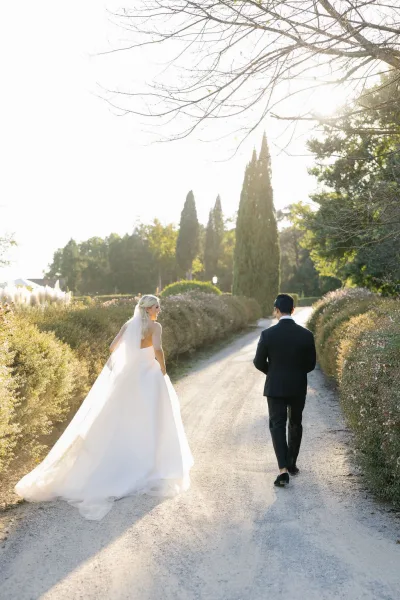 First look moment as bride walks to groom in a wedding dress and long veil, with tuxedo suit, on a sunlit garden path lined with tall trees