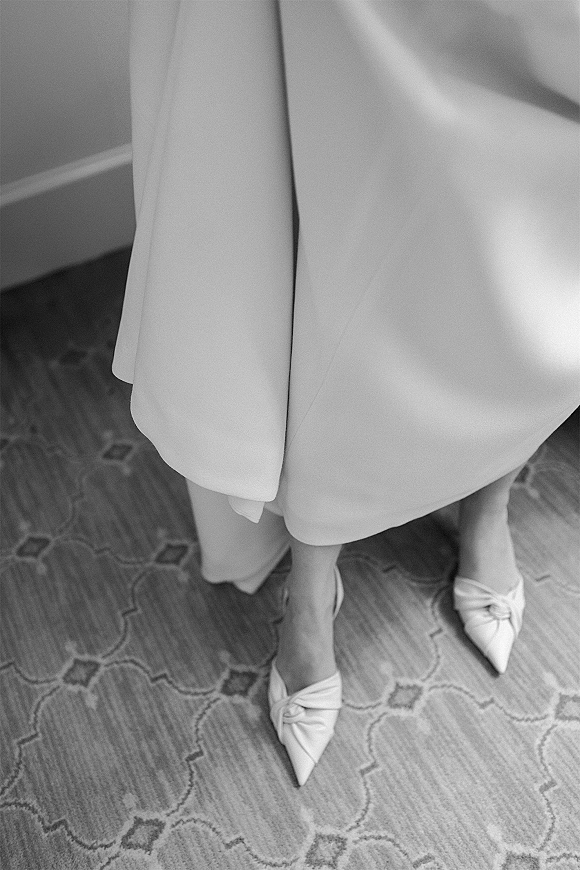 Bridal shoes, white pointed toe heels with knot detail resting on patterned carpet beside the bridal dress hem and baseboard trim