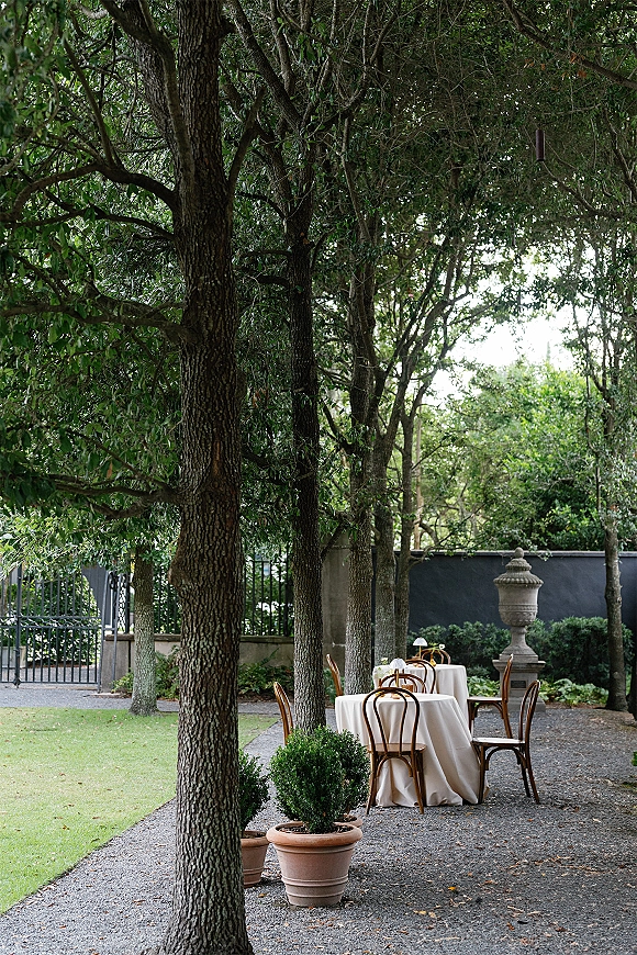 Outdoor reception setup with garden reception tables, round white-clothed settings, bentwood chairs and table lamps on a tree-shaded lawn