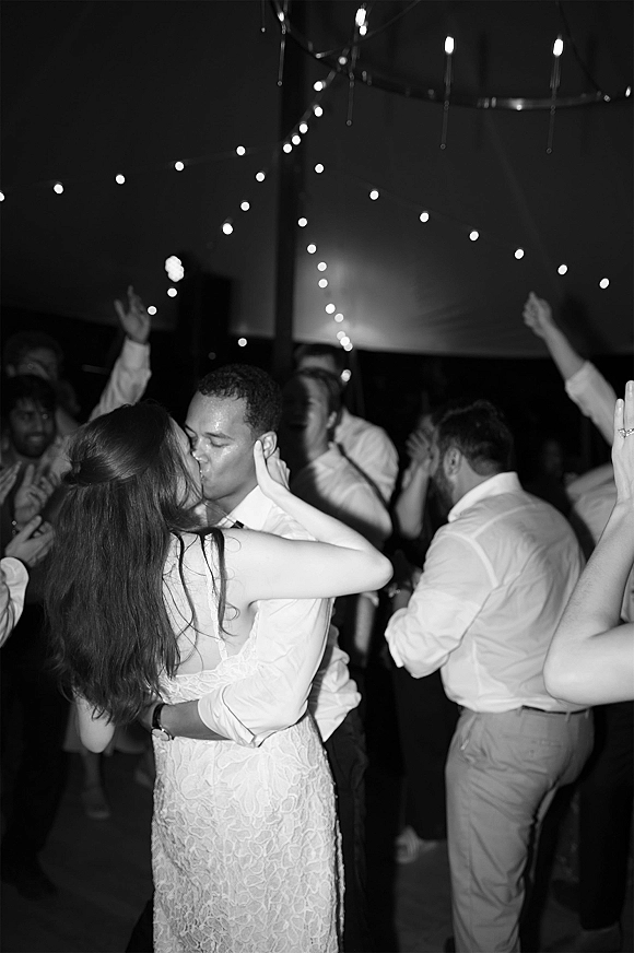 Wedding kiss during a last dance on a crowded reception dance floor under string lights, bride in lace dress as guests cheer in a tent
