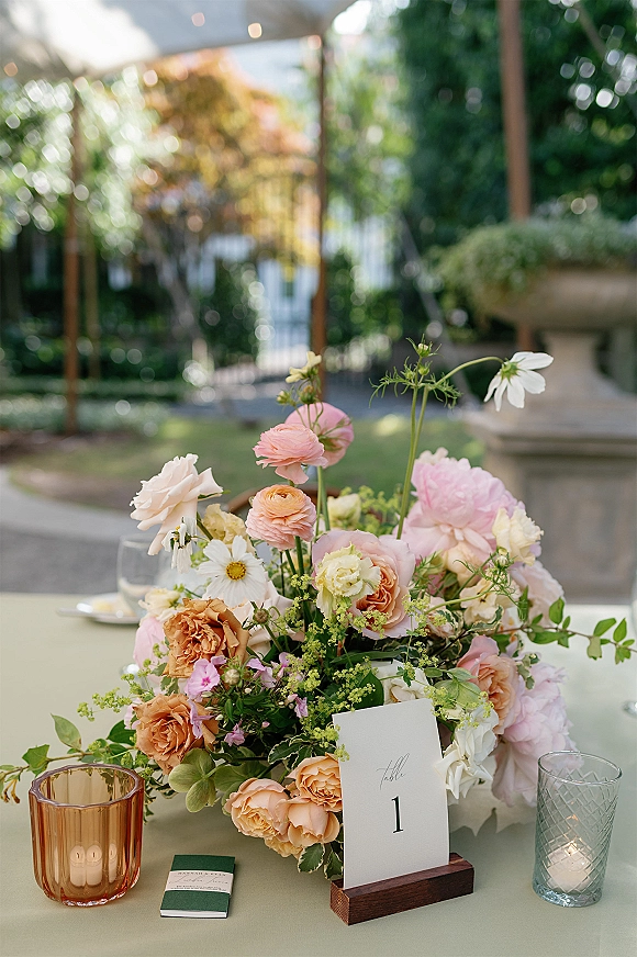Reception centerpiece with roses and ranunculus, greenery, table number card, and amber votives on a garden patio near a stone fountain