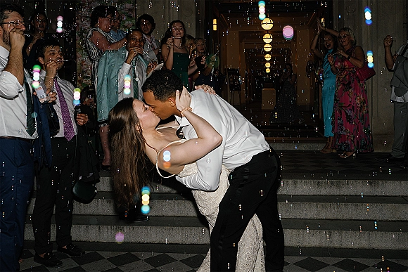 Wedding kiss in a wedding dip kiss as groom lifts bride on stone steps, guests cheer and blow bubbles under pendant lights with champagne glasses