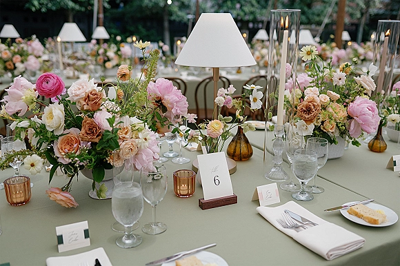 Reception tablescape with floral centerpieces, taper candles, and table lamps on sage linens in an outdoor garden under string lights