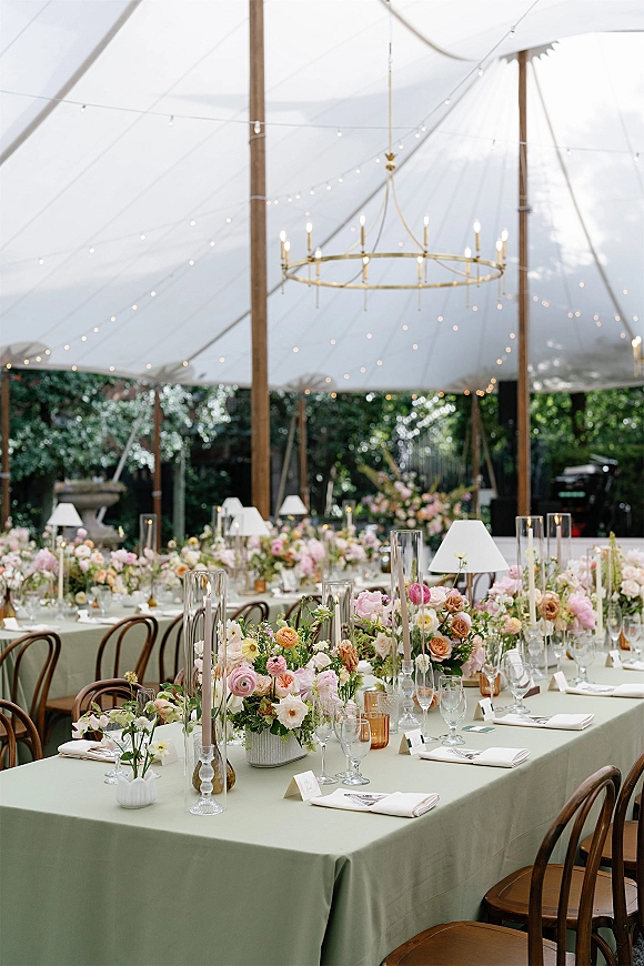 Reception tablescape with long banquet table decor in sage green linens, pastel rose centerpieces, taper candles, and string lights under a tent at dusk