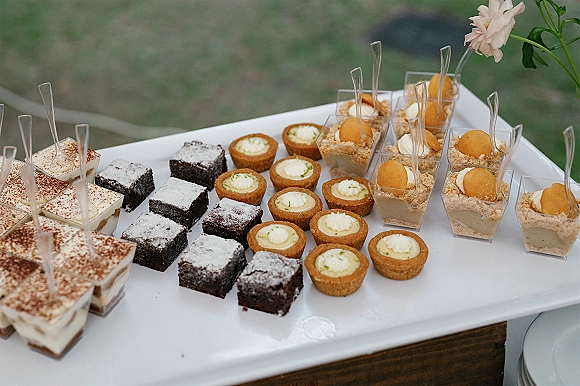 Wedding dessert table with mini dessert cups and spoons, brownie bites and mini tarts on a white platter atop a wooden table in outdoor greenery