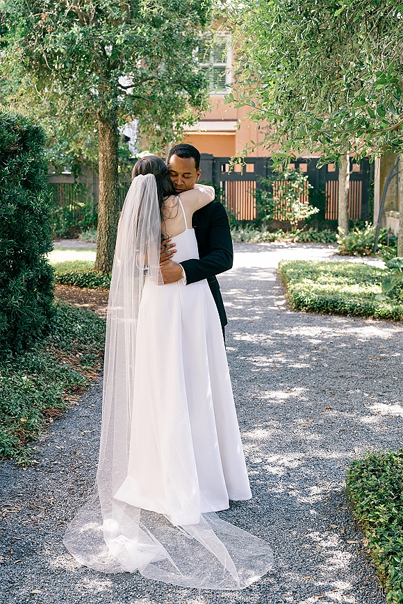 Wedding couple portrait of bride and groom hug on a tree-lined garden path, her long veil trailing over a strapless gown beside him in a black suit