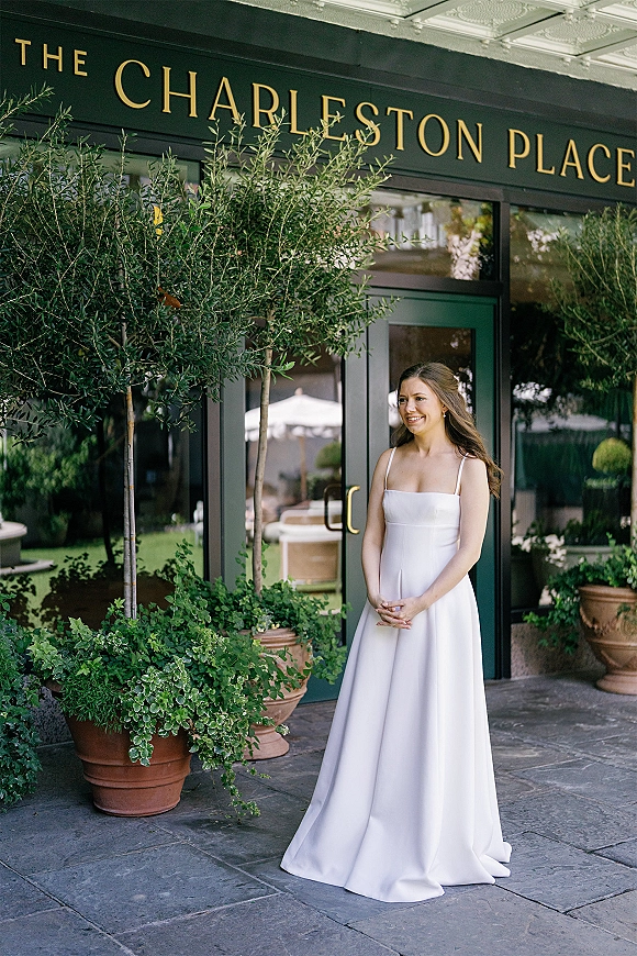 Bridal portrait of a bride in a simple wedding dress with spaghetti straps, hands clasped by glass doors and terracotta planters