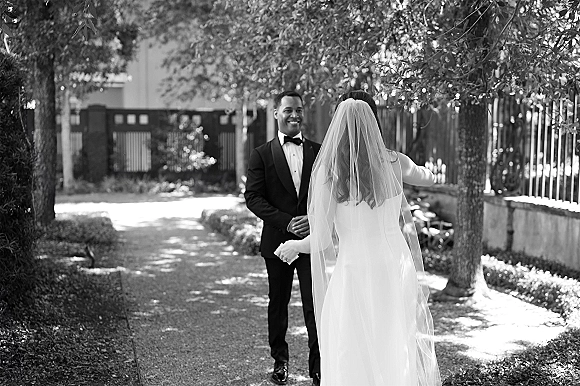 Wedding first look as the bride in a long sleeve dress and veil approaches her groom in a tuxedo on a dappled garden path