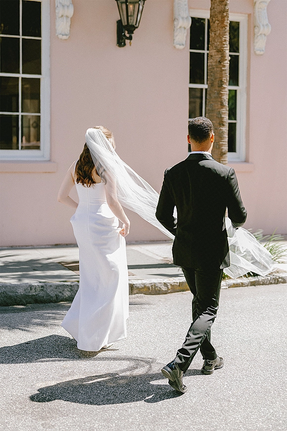 Wedding couple portrait of bride and groom walking away, long veil blowing behind them along a pink stucco wall on a sunny street