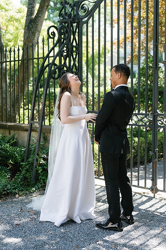 Couple portrait of bride and groom laughing, holding hands in front of a wrought iron gate, her veil and dress train flowing
