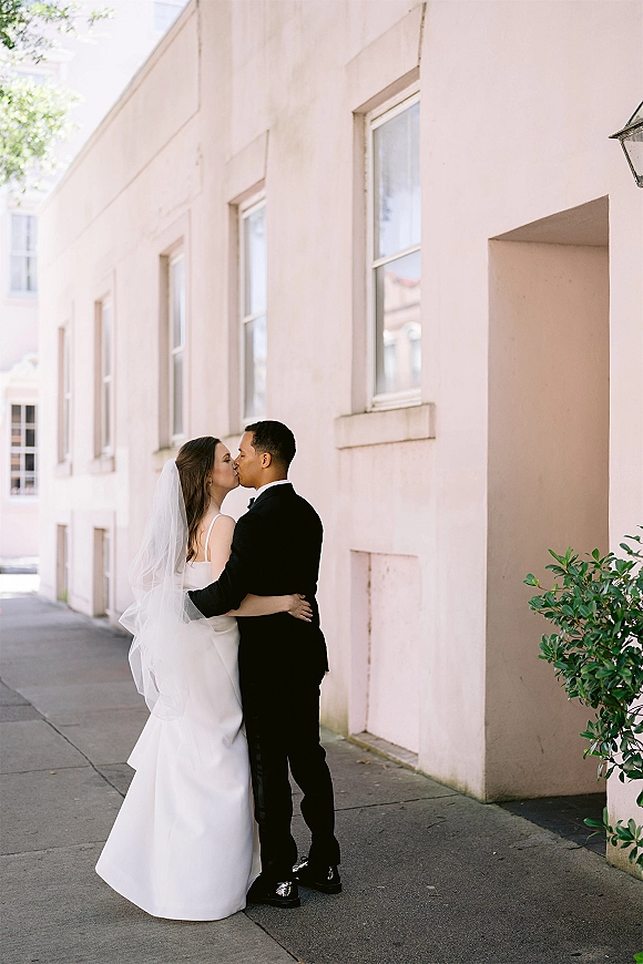 Wedding kiss as bride and groom kiss on a city sidewalk, her long veil trailing behind his tuxedo by a pastel building facade
