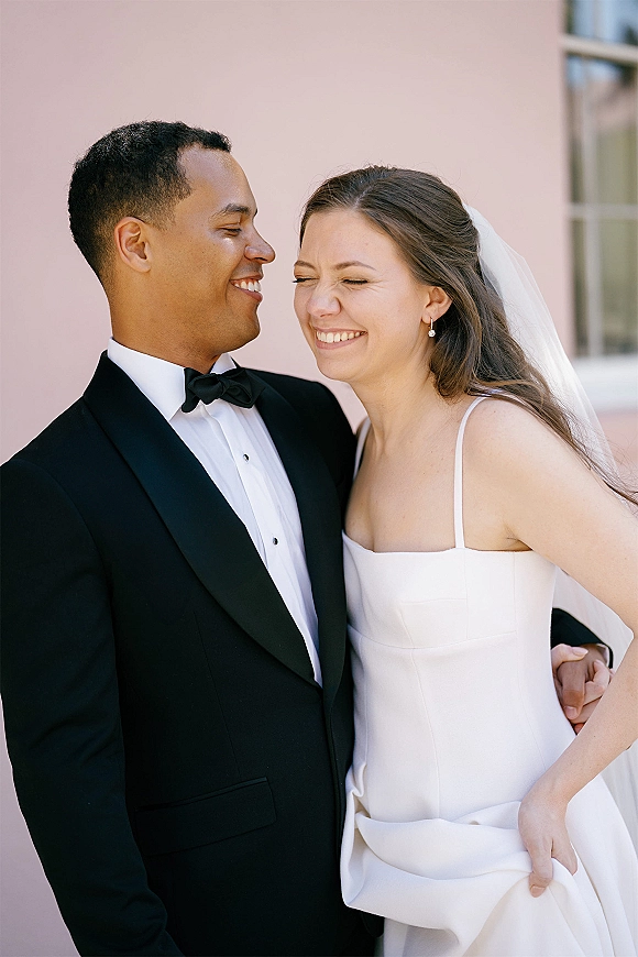Couple portrait of bride and groom laughing, her veil over a strapless gown as he smiles in a tuxedo by a pink wall and window light