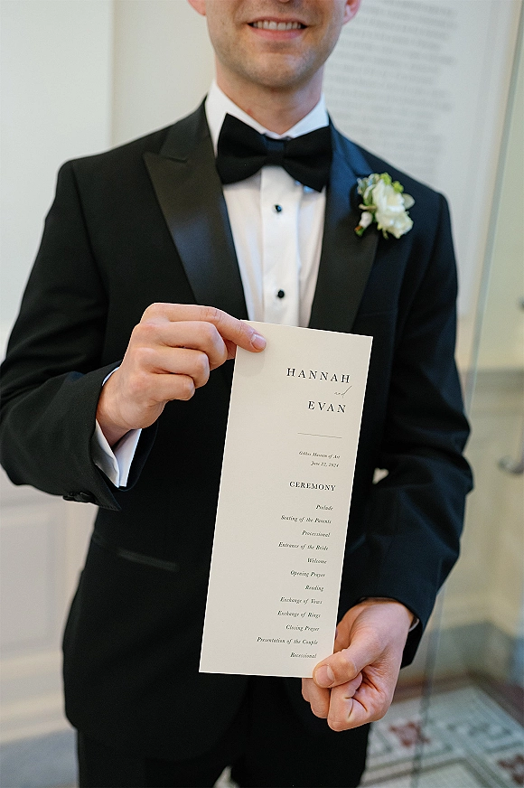 Wedding ceremony program held by a groom in a tuxedo with black bow tie and boutonniere, standing indoors by framed artwork