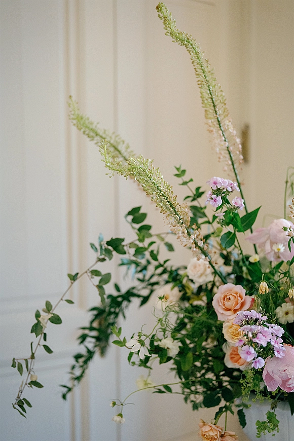 Wedding floral arrangement wedding flower centerpiece with peach roses, peonies, pampas grass, and trailing greenery against a cream paneled wall