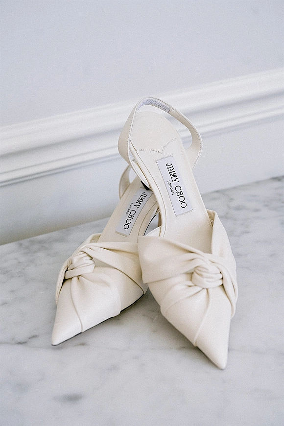 Bridal shoes in ivory wedding shoes style, slingback pointed-toe heels with a knotted detail arranged on a marble surface against a white wall