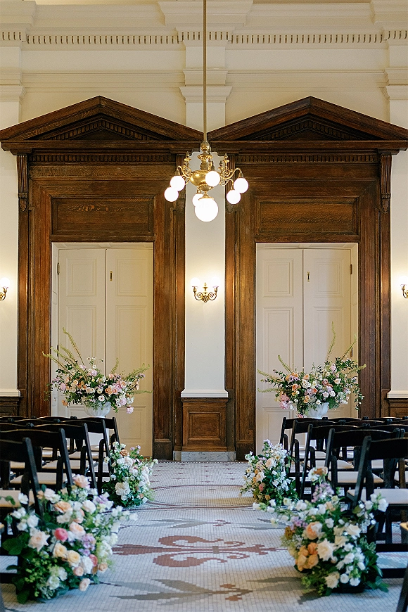 Ceremony aisle decor with floral aisle markers and greenery lining a mosaic tile aisle, framed by black folding chairs in a wood-paneled hall