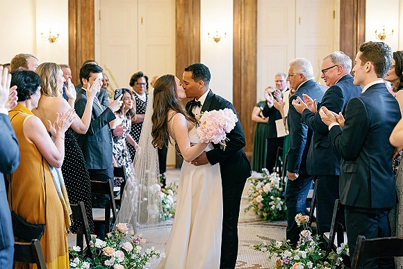 Wedding kiss during the ceremony recessional kiss as bride in veil and groom in tux hold a pink bouquet, guests stand by chairs indoors