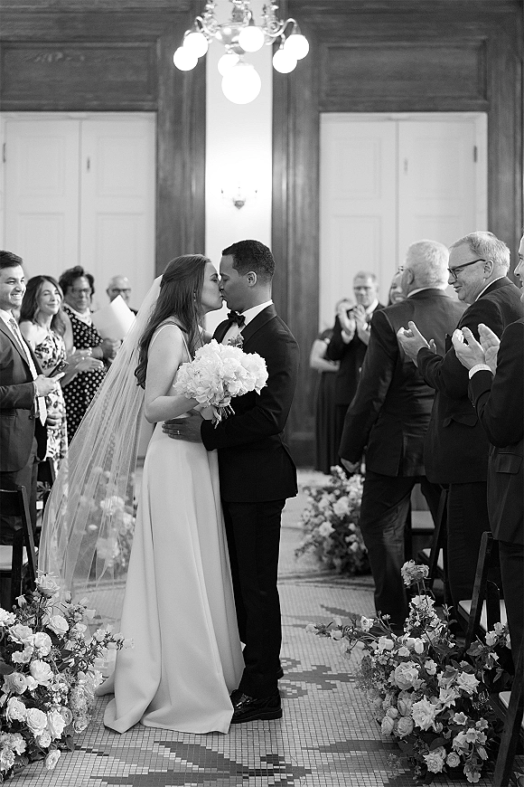 Wedding kiss as newlyweds embrace in the aisle, bride in veil holding bouquet beside groom in tuxedo with guests in wood-paneled room