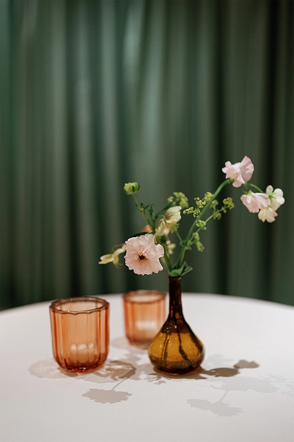 Reception centerpiece with a bud vase of pink flowers and amber glass vase beside amber votive holders on a round table, green drape backdrop
