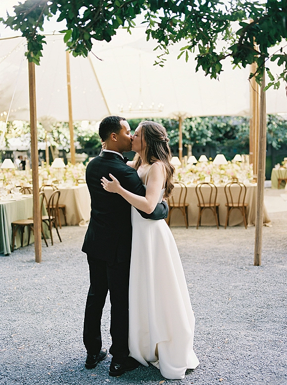Wedding kiss as bride and groom kissing under a tent canopy, string lights glowing above long reception tables and ivy wall backdrop