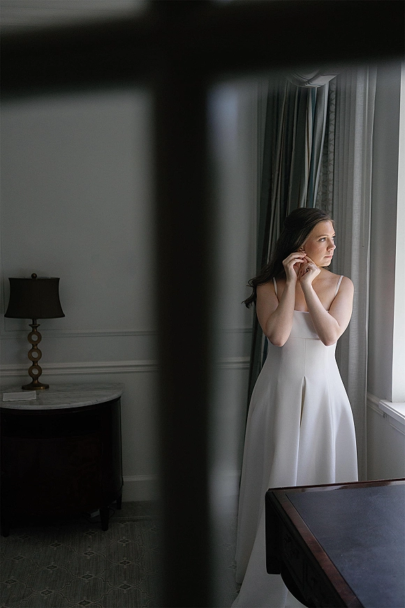 Bridal portrait of a bride putting on earrings in a simple satin wedding dress by a window with sheer curtains in a hotel suite