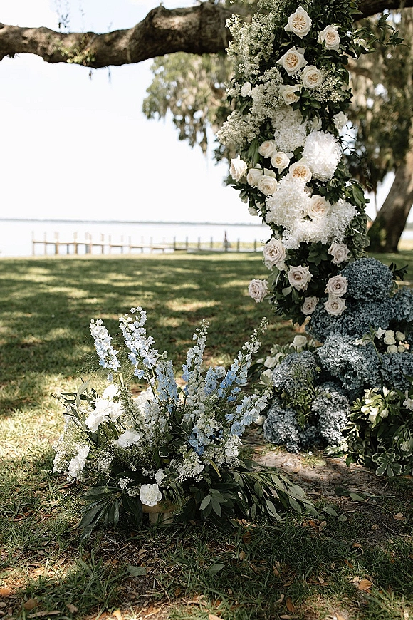 Wedding ceremony flowers on a floral arch installation with white roses and blue hydrangeas under an oak tree by the waterfront dock